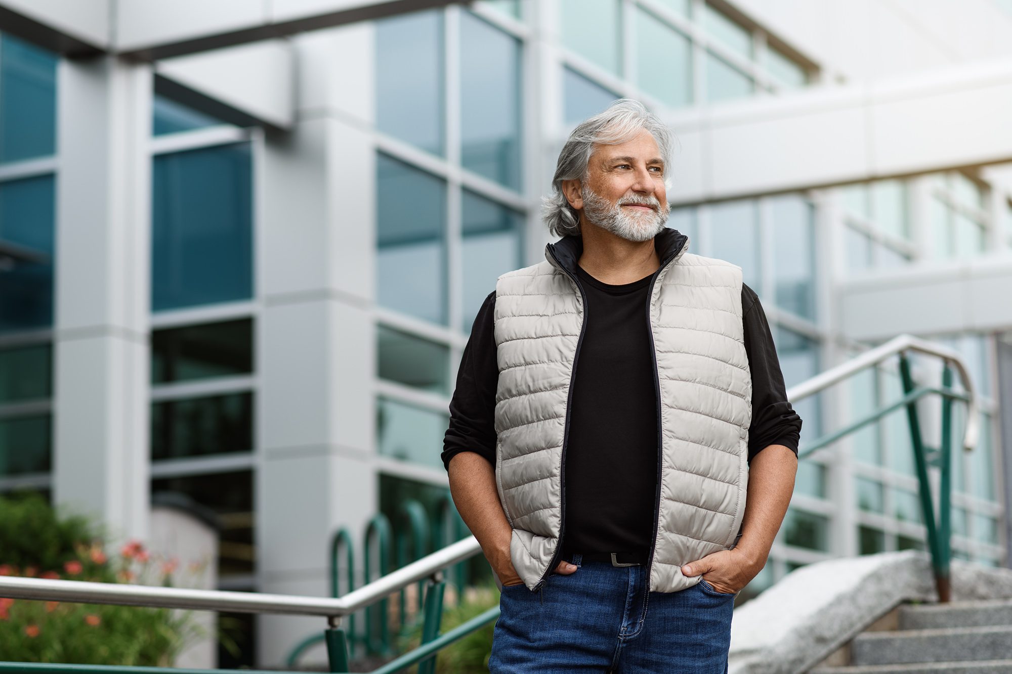 Portrait of a white hair and bearded man wearing a white vest, in front of a modern architectural building.