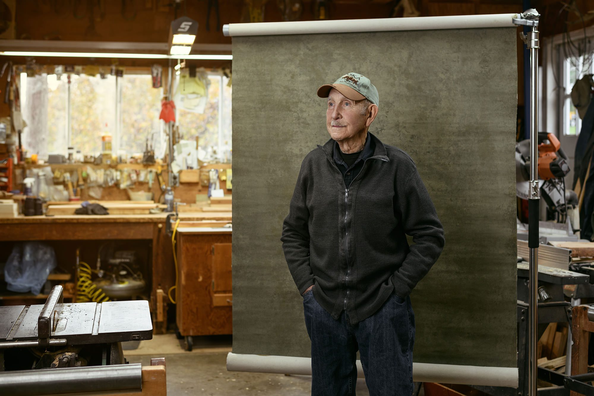 Environmental portrait of senior man in his wood workshop, looking off camera. A hand painted canvas backdrop is behind him.