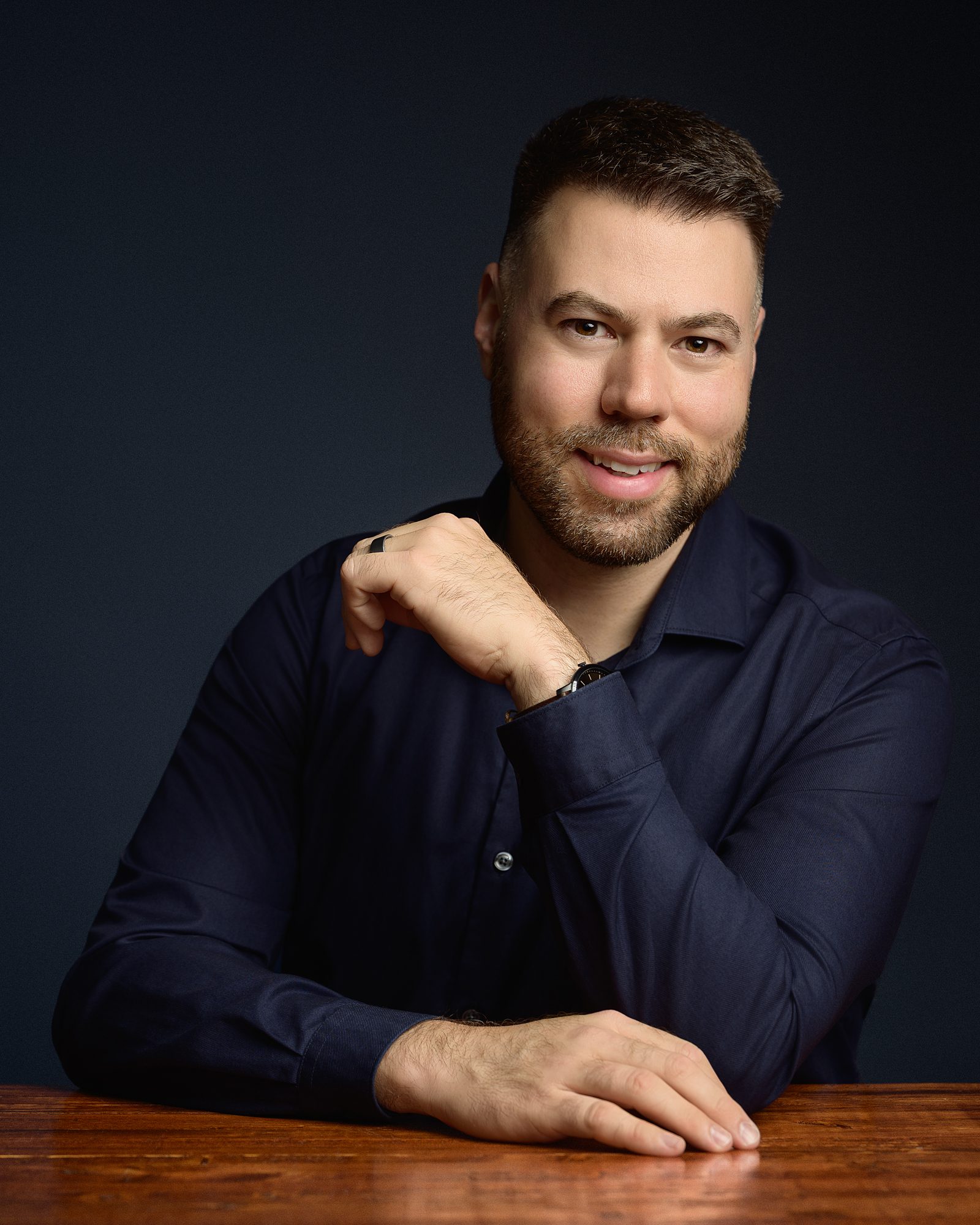 Portrait of Philip Boudreau, sitting at a wood table, blue background.